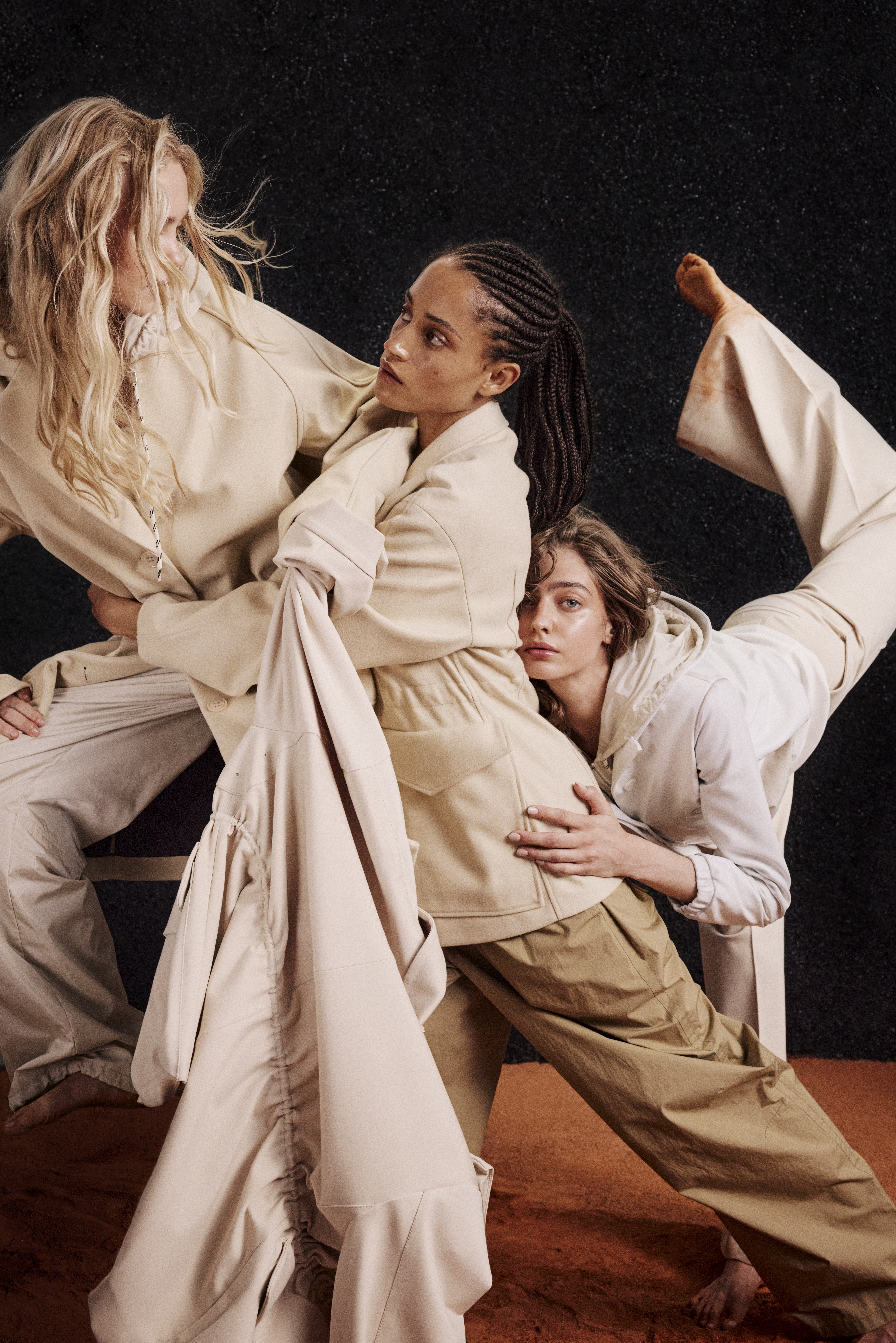 three women wearing white and beige clothes like coats and jackets and wideleg pants, dancing on red sand and in front of a black background