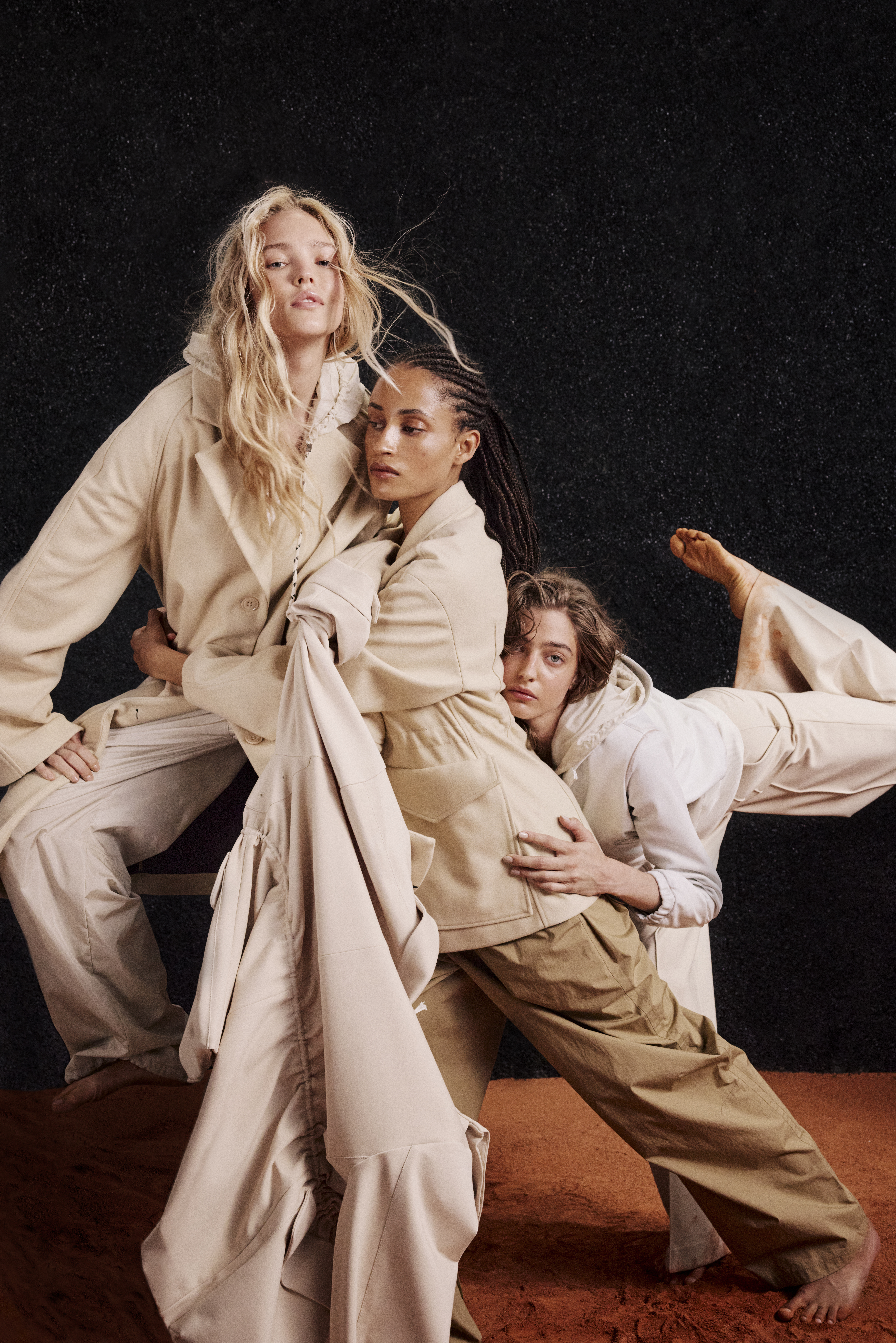 three women wearing white and beige clothes like coats and jackets and wideleg pants, dancing on red sand and in front of a black background