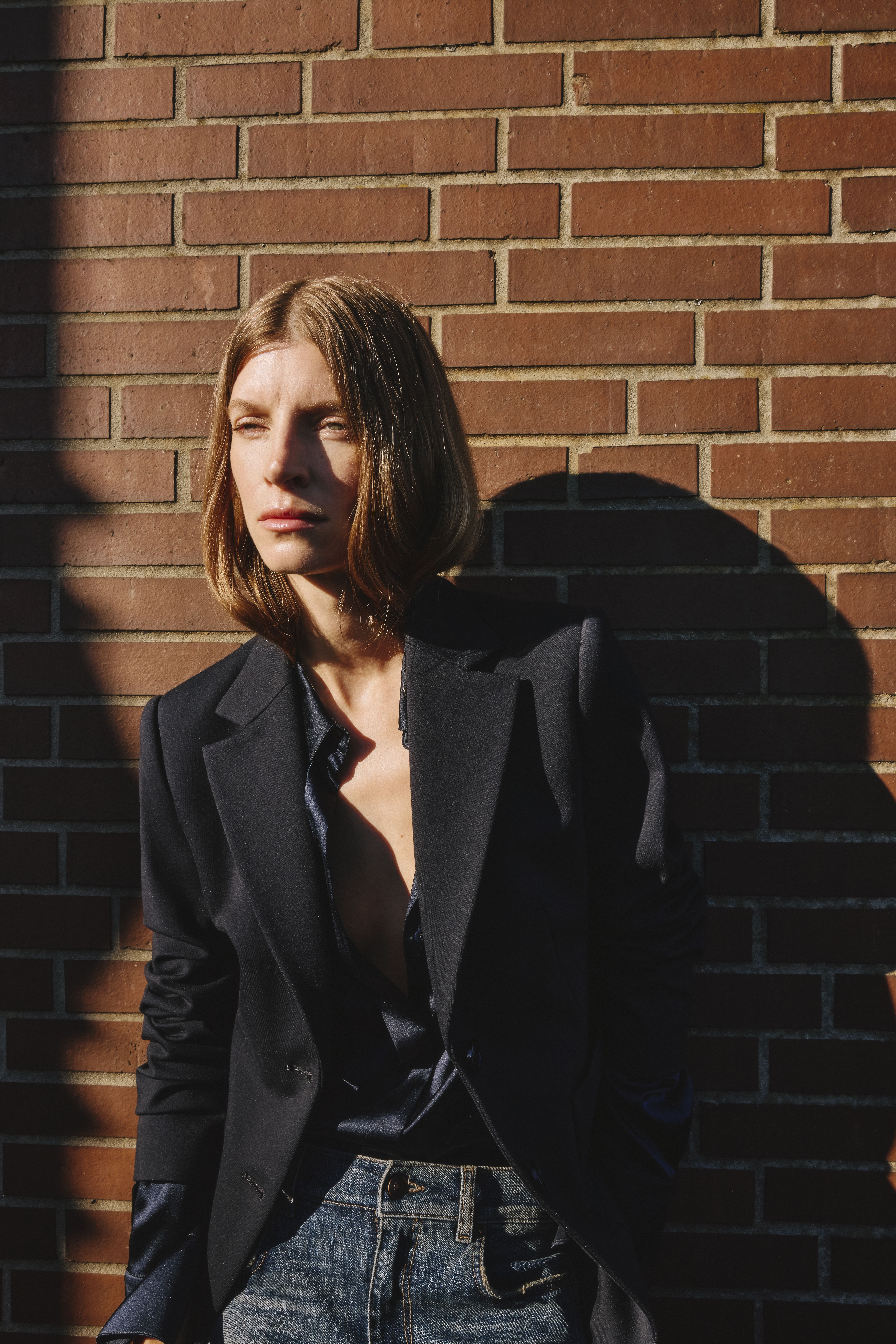 A woman with a black blazer, a blue blouse and a blue jeans, leaning on a wall