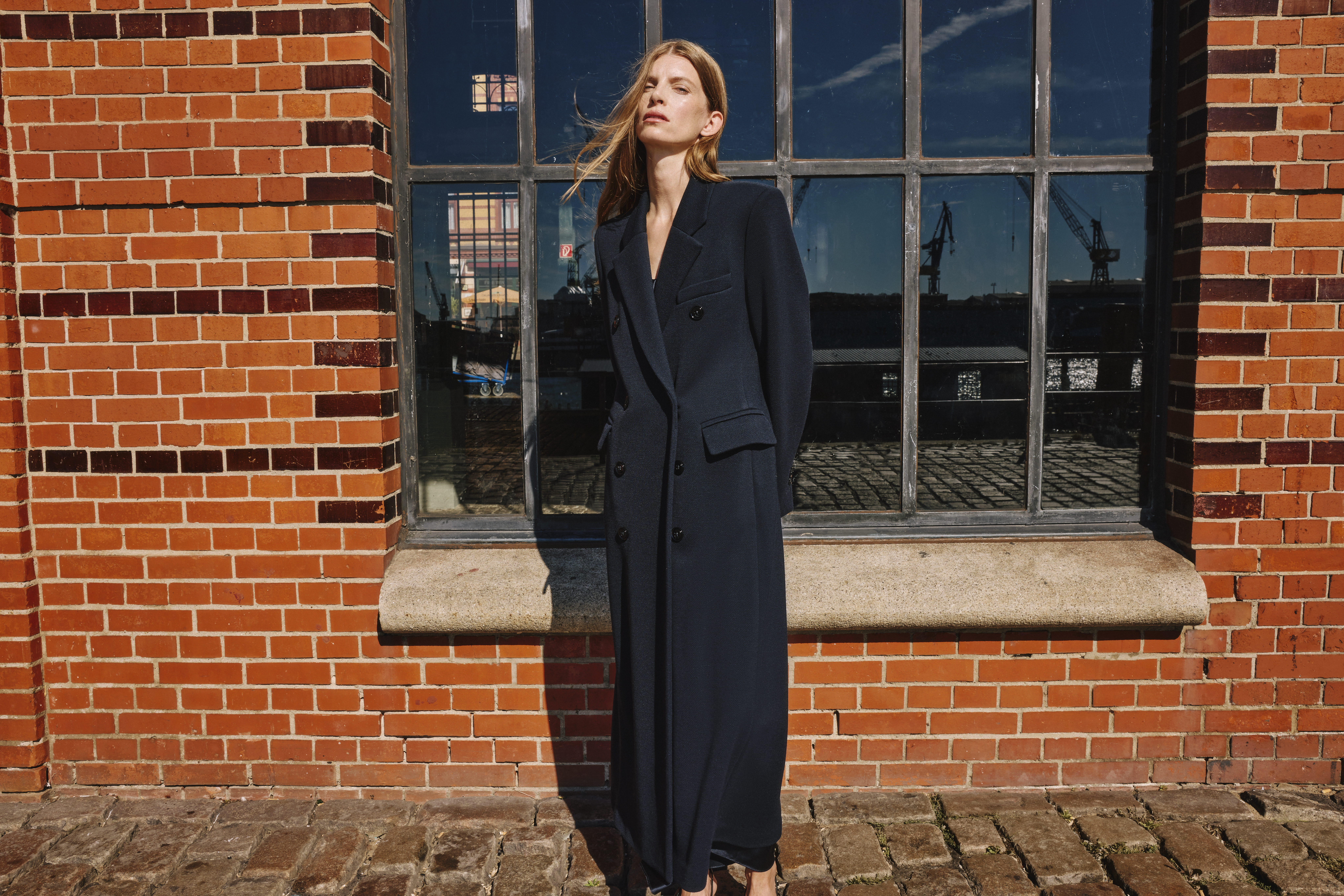 woman standing in a blue long coat in front of a big window