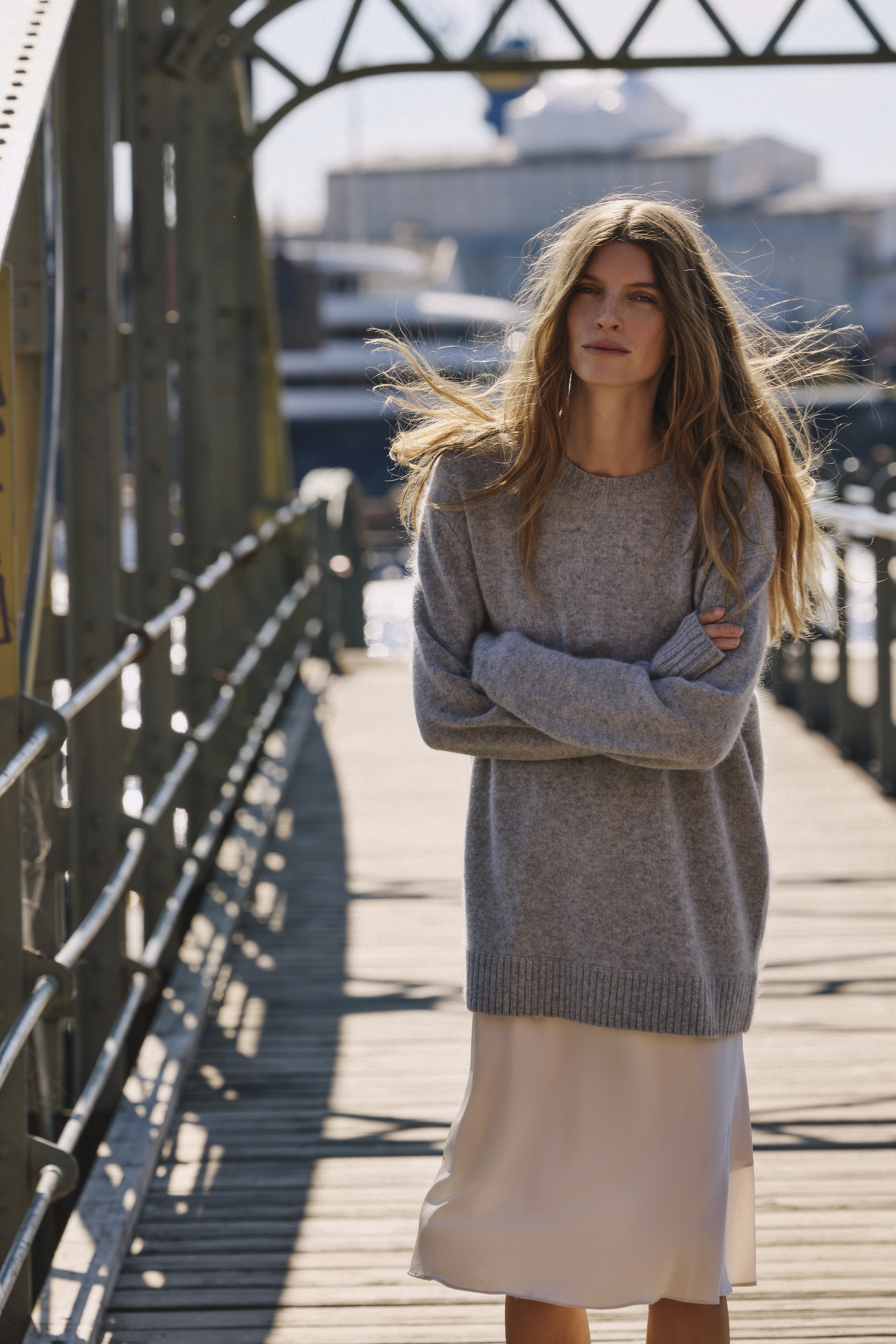 the woman is wearing a grey pullover and a white satin skirt on a bridge