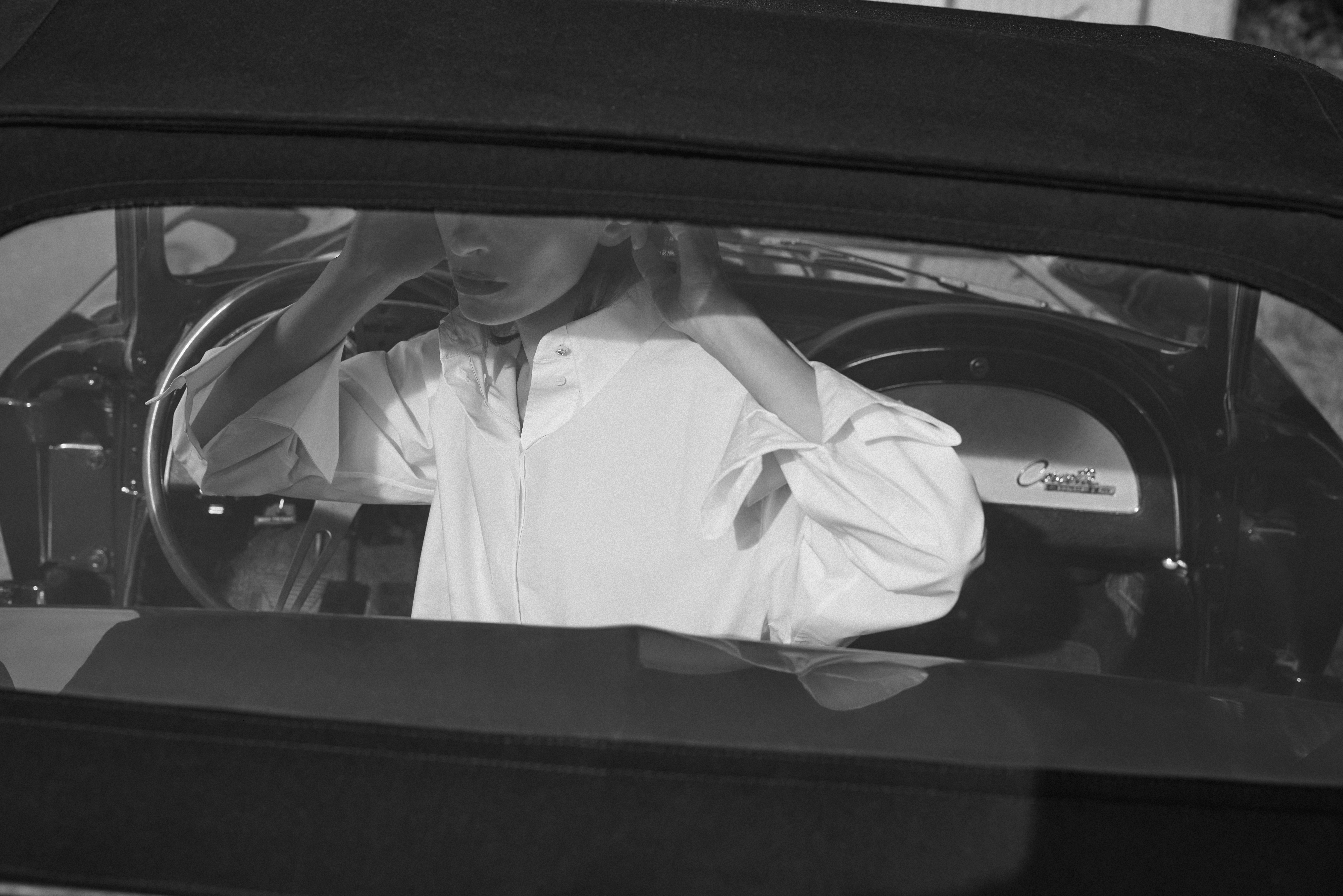 a woman is sitting in a car wearing a white cotton blouse putting her hair behind her ears