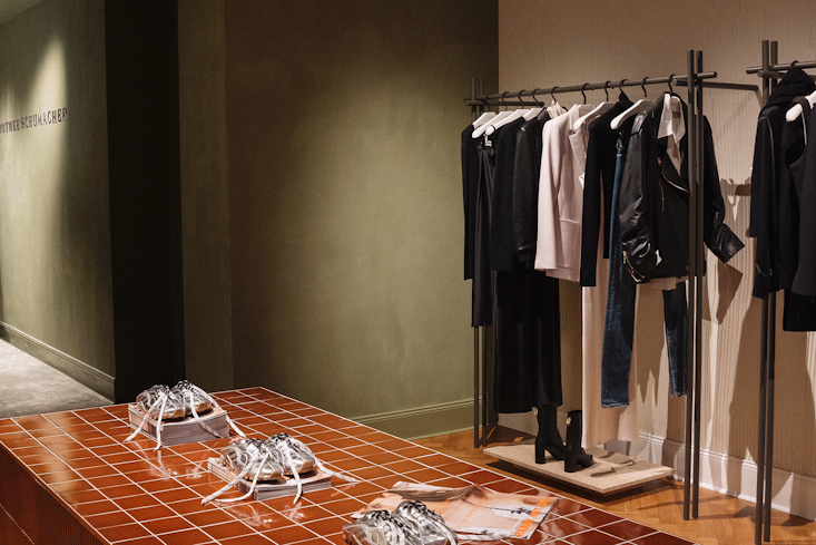 Innenansicht des Dorothee Schumacher Geschäfts mit Kleiderständern voller Damenbekleidung und einem gefliesten Präsentationstisch.