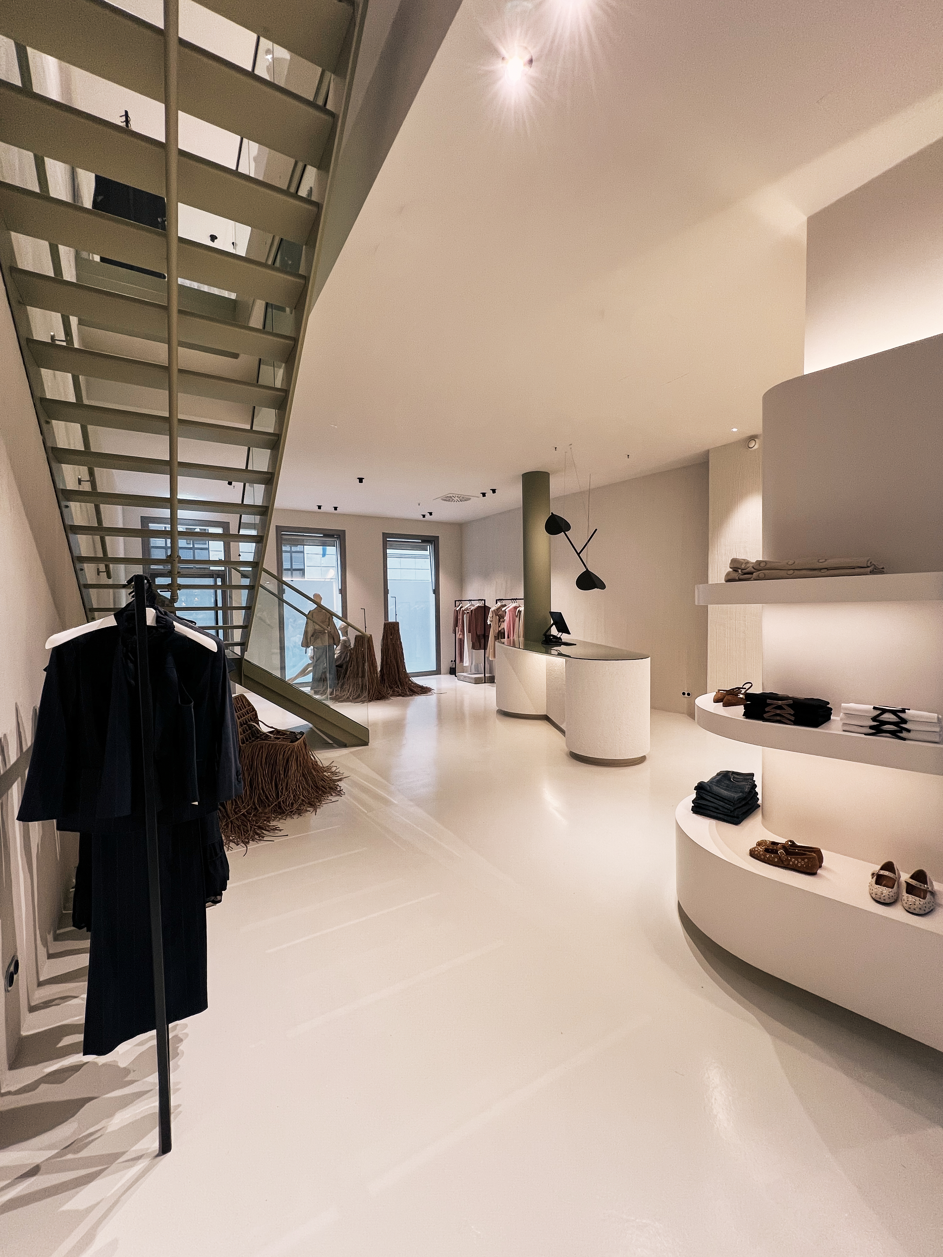 Blick in den Dorothee Schumacher Store unter einer minimalistischen Treppe, mit Kleiderstangen und runden Präsentationsmodulen für Schuhe und Accessoires.