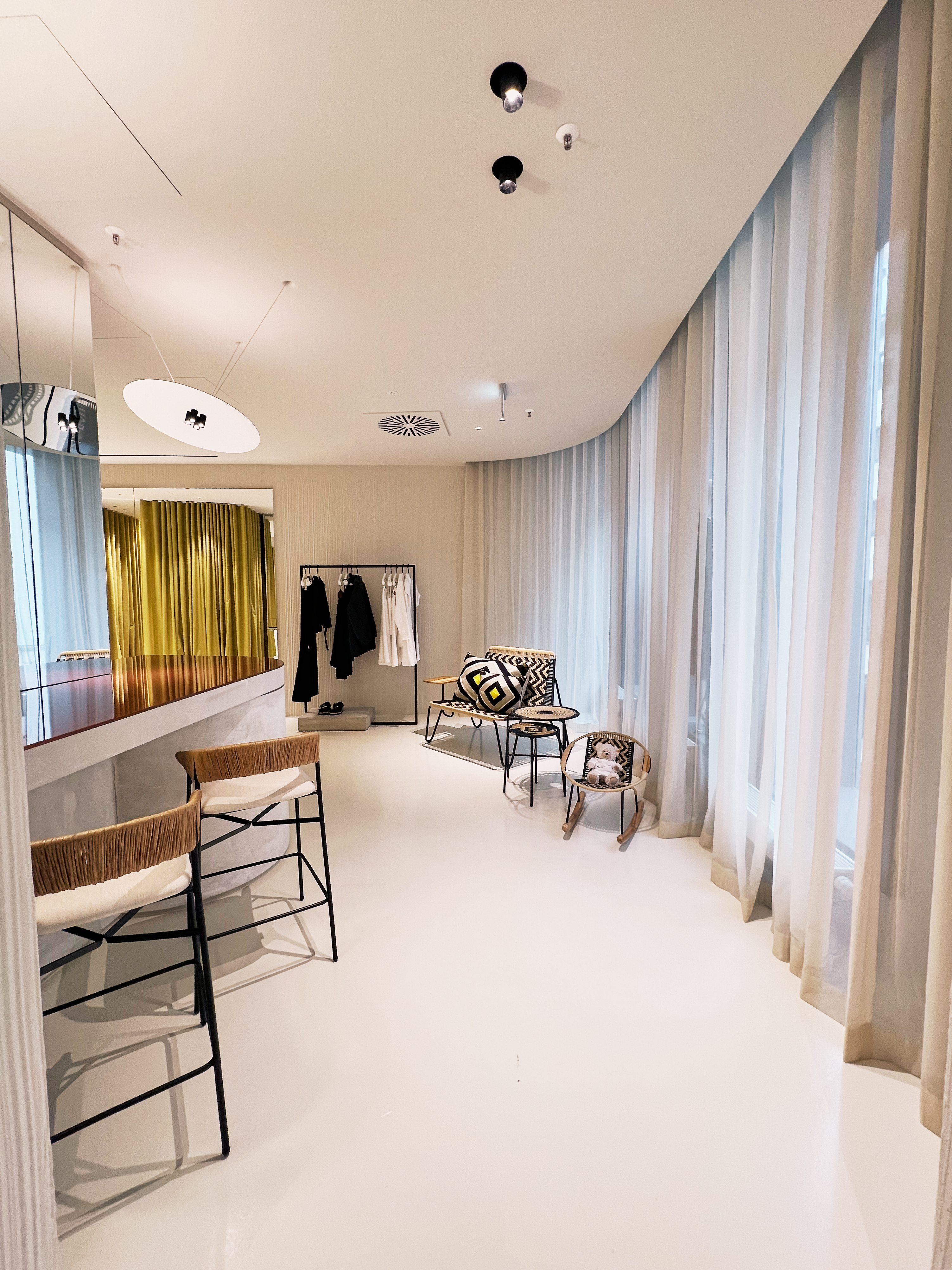 Stilvoller Lounge-Bereich im Dorothee Schumacher Store mit geflochtenen Barhockern, einer geschwungenen Theke und bodentiefen weißen Vorhängen für eine luftige Atmosphäre.