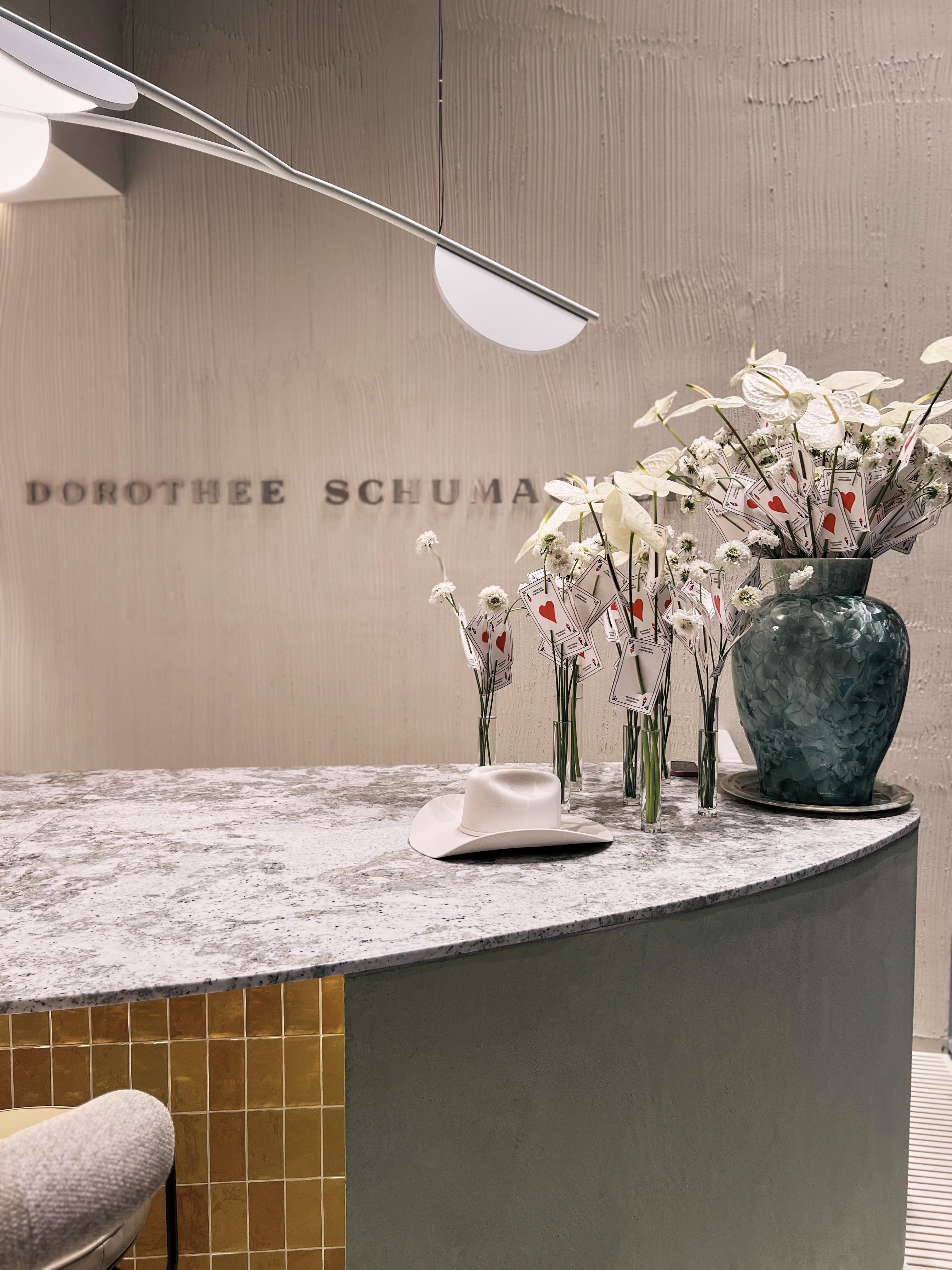 Close-up of a marble counter with the Dorothee Schumacher brand lettering in the background, decorated with white flowers and a white hat.