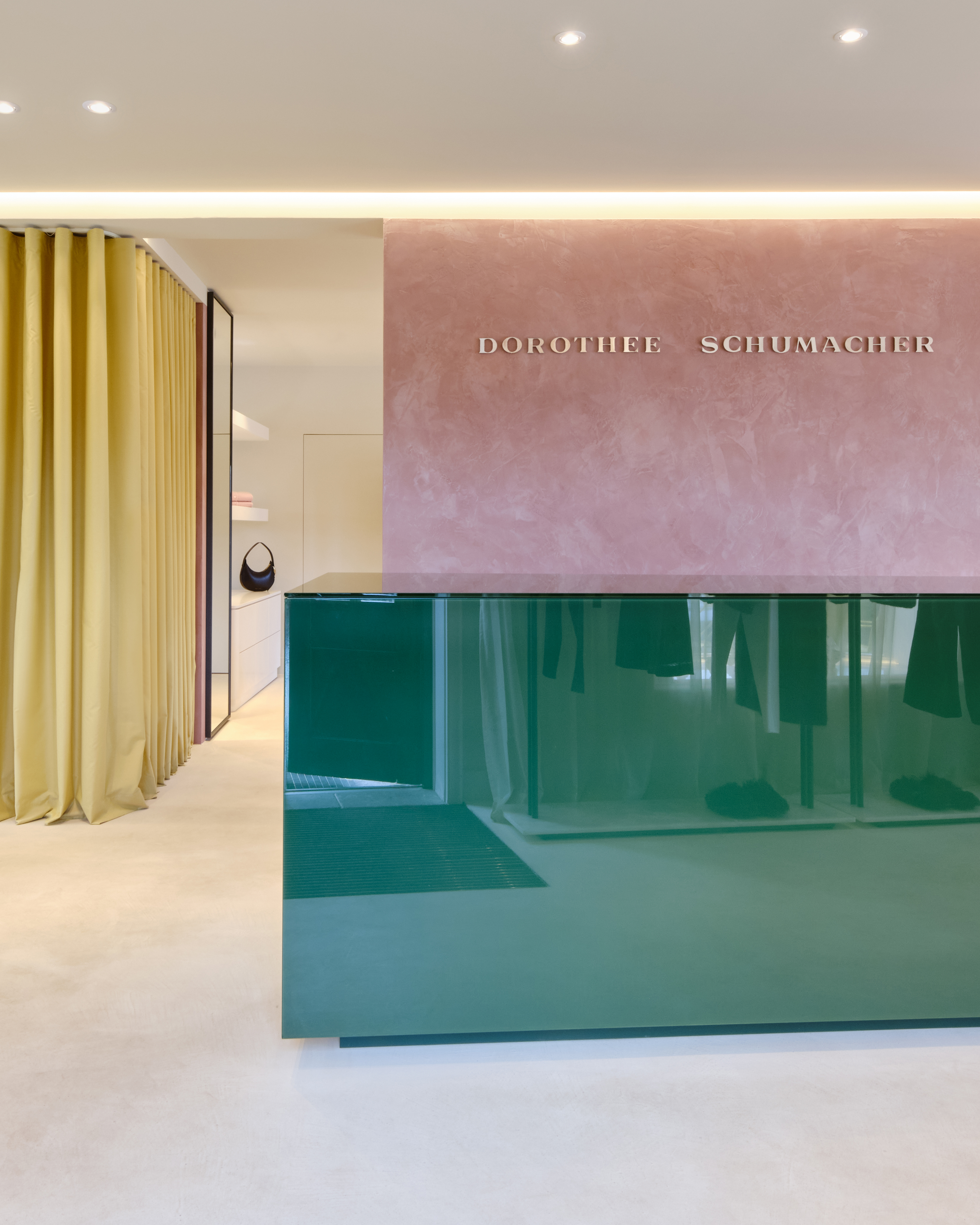 Moderner Empfangsbereich im Dorothee Schumacher Geschäft mit einer glänzenden grünen Theke, einer rosa Wand mit dem 'DOROTHEE SCHUMACHER' Logo und gelben Vorhängen.